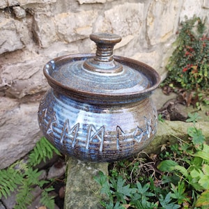 Op de afbeelding: Een ronde keramische pot met deksel, voorzien van een glanzend glazuur in tinten diepblauw en aards bruin. Het buitenoppervlak is versierd met een band van gegraveerde runensymbolen die worden geaccentueerd door de blauwe glazuurafwerking.