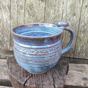 Può includere: Una tazza in ceramica fatta a mano con smalto blu e marrone e una superficie ruvida. La tazza presenta un corpo arrotondato, un bordo largo e un manico curvo con un piccolo poggia-pollice sulla parte superiore. L'esterno mostra striature.