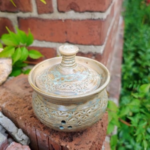 Könnte beinhalten: Ein runder, beige-blauer Keramik-Knoblauchtopf mit abnehmbarem Deckel. Das Gefäß verfügt über ein strukturiertes, gestempeltes Blumenmuster auf dem Körper und dem Deckel, einen kleinen Knopfgriff und drei kreisförmige Belüftungslöcher am Boden.