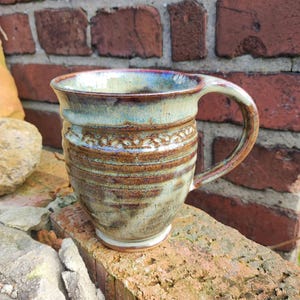 Puede incluir: Taza de cerámica hecha a mano con un diseño único. La taza presenta un esmalte azul claro y marrón con una banda decorativa en el medio. El asa es robusta y bien integrada. La taza está colocada contra una pared de ladrillos.