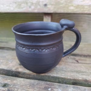 Könnte beinhalten: Eine schwarze Keramiktasse mit abgerundetem Körper und einem kleinen Henkel. Die Tasse hat ein dekoratives Design am oberen Rand.