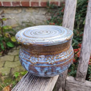Può includere: Un burriera in ceramica rotonda con coperchio, caratterizzata da una smaltatura maculata blu e marrone. Il coperchio presenta un motivo a spirale e il corpo è decorato con un rilievo floreale ripetuto nella parte superiore, dal finish rustico.