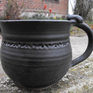 Könnte beinhalten: Eine handgefertigte schwarze Keramiktasse mit rundem Boden und großem Henkel. Die Tasse hat ein dekoratives Band aus geschnitzten Mustern in der Mitte. Die Tasse eignet sich für Kaffee, Tee oder andere Getränke.