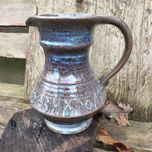 Könnte beinhalten: Handgefertigter Keramikkrug mit blau-brauner Glasur. Der Krug hat einen Henkel und einen Ausguss. Der Boden des Kruges weist geschnitzte Runen auf. Der Krug steht auf einer Holzoberfläche.