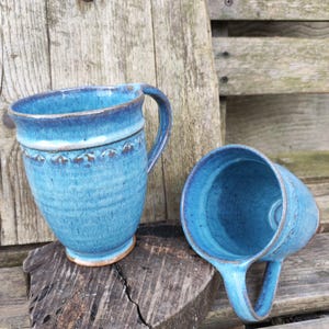 Può includere: Due tazze in ceramica fatte a mano con una vivace glassa turchese. Una tazza è in posizione verticale, l'altra è di lato. Entrambe le tazze hanno un manico e una fascia decorativa vicino al bordo. Le tazze sono appoggiate su una superficie di legno stagionato.