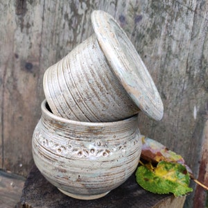 Może przedstawiać: Zestaw dwóch ceramicznych pojemników na masło z białą glazurą i brązowymi smugami. Pojemniki są ułożone jeden na drugim, z pokrywką na górze. Pojemniki mają rustykalny, ręcznie robiony wygląd.
