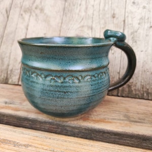 Könnte beinhalten: Eine tealblaue Keramiktasse mit einem einzigartigen Henkel und einem dekorativen Design am Rand. Die Tasse steht auf einer Holzoberfläche.