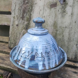 Peut inclure: Un beurrier en céramique bleu avec un couvercle. Le couvercle a un bouton sur le dessus et le plat a des runes gravées sur le côté.