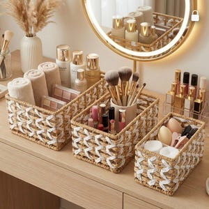 Op de afbeelding: Een kaptafelopstelling met geweven manden met schoonheidsproducten. De manden bevatten make-up borstels, lippenstiften, huidverzorgingsflessen en opgerolde handdoeken. Een verlichte spiegel en een vaas met gedroogde planten zijn ook aanwezig.
