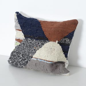 May include: A square decorative pillow with a patchwork design. The pillow features various textured fabrics in shades of navy blue, brown, gray, and cream. The pillow is approximately 18 inches square.