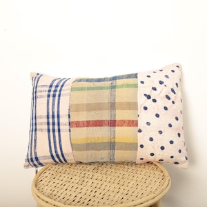 Peut inclure: Coussin rectangulaire décoratif avec un motif patchwork. Le coussin présente des sections de tissu à rayures bleues, un motif à carreaux avec des rayures rouges et jaunes, et un tissu rose à pois bleu marine. Il est posé sur un tabouret en osier.