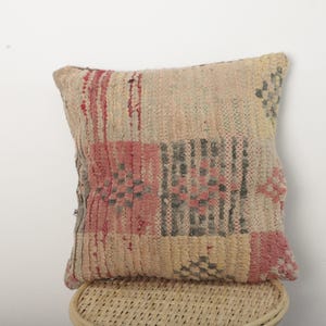 Peut inclure: Un coussin carré texturé avec un motif patchwork. Le coussin présente un mélange de couleurs beige, rouge et gris, disposées en motifs géométriques. Il repose sur une surface tissée de couleur claire, suggérant un style rustique ou bohème.