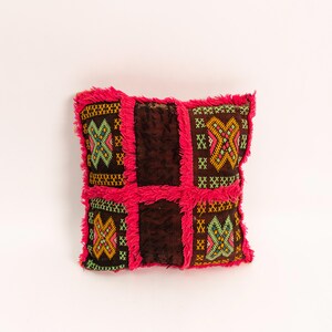 Peut inclure: Coussin décoratif carré avec un centre marron et des motifs géométriques brodés en jaune, vert et rose. Le coussin a une bordure à franges rose vif, ajoutant une touche de couleur et de texture au design.