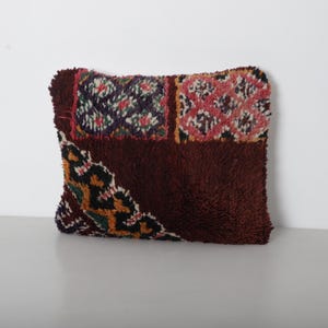 Peut inclure: Un coussin décoratif rectangulaire avec une surface texturée et hirsute. Le coussin présente un motif patchwork avec des sections bordeaux foncé, des carrés à motifs roses, violets et verts, et un coin avec un motif noir, orange et jaune.