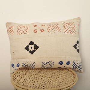 Peut inclure: Coussin rectangulaire crème avec des motifs géométriques rouges, noirs et bleus. Le coussin présente une texture tissée et est posé sur un tabouret en osier. Le design comprend des motifs de losanges et floraux.