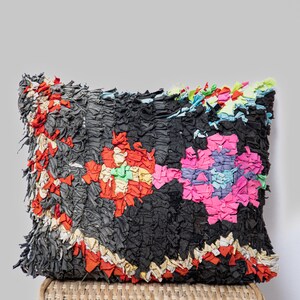 Peut inclure: Un coussin rectangulaire noir avec des motifs floraux colorés. Le coussin est fait de bandes de tissu dans les tons de rouge, rose, jaune et bleu. Le coussin est posé sur une surface tissée.