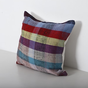 Peut inclure: Un coussin carré avec un motif à carreaux coloré. Le coussin présente des rayures rouges, violettes, bleues et jaunes. Le coussin est posé sur une surface blanche.