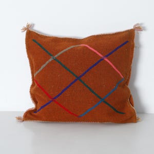 Peut inclure: Un coussin carré de couleur rouille avec un motif de grille colorée cousue. La grille est composée de lignes entrecroisées rouges, bleues, vertes, roses et grises. Le coussin a de petits pompons à chaque coin, et est un élément de décoration intérieure.
