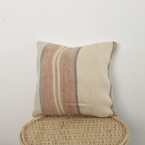 Peut inclure: Coussin carré tissé avec un fond crème et des rayures verticales dans les tons de marron, gris et bleu. Le coussin est posé sur une chaise en osier tressé.