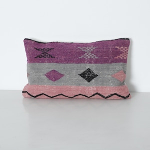 Peut inclure: Un coussin rectangulaire décoratif avec un motif tissé. Le coussin présente des bandes de violet, de gris et de rose, avec des motifs géométriques noirs. Les bords du coussin sont soulignés en noir, ajoutant un détail contrastant.