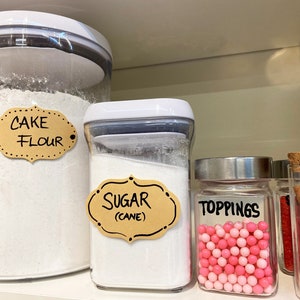May include: Three clear glass jars with white lids on a white shelf. The jars are labeled with brown paper labels and black marker. The labels read "Cake Flour", "Sugar (Cane)", and "Toppings". The jar labeled "Toppings" contains pink and white sprinkles.