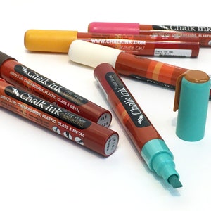 Peut inclure: Gros plan sur un ensemble de marqueurs à encre de craie multicolores. Les marqueurs sont étiquetés "Chalk Ink" et "Hi-Rise Markers". Le texte sur les marqueurs indique "Writes on: Chalkboards, Plastic, Glass & Metal".