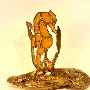 May include: A stained glass seahorse sculpture in shades of amber and gold, with green accents, mounted on a piece of driftwood. Small pebbles are at the base of the seahorse. The seahorse is approximately 6 inches tall.