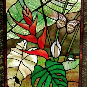 May include: Stained glass panel depicting a tropical scene. Features red flowers, green leaves, butterflies, and a small frog. The artwork is framed with a dark border and showcases vibrant colors and intricate details.
