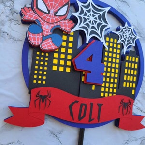 May include: A red and blue Spiderman cake topper with a black and yellow cityscape, a blue number 4, and a red banner with the name "Colt" and a black Spiderman logo.