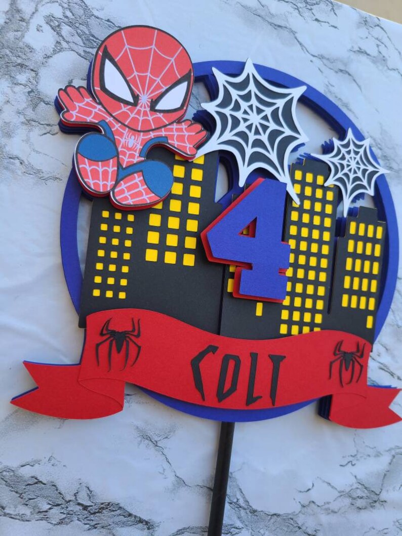 May include: A red and blue Spiderman cake topper with a black and yellow cityscape, a blue number 4, and a red banner with the name "Colt" and a black Spiderman logo.