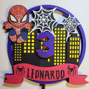 May include: A red and blue Spiderman cake topper with a purple number 3 and a red banner that says "LEONARDO". The topper features a cityscape with yellow windows and black spiderwebs.