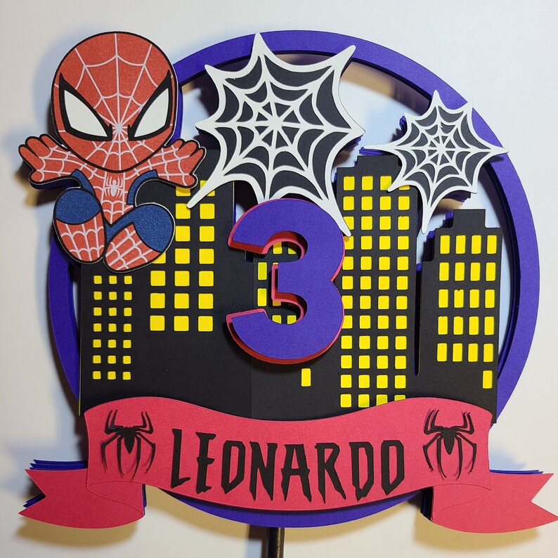 May include: A red and blue Spiderman cake topper with a purple number 3 and a red banner that says "LEONARDO". The topper features a cityscape with yellow windows and black spiderwebs.