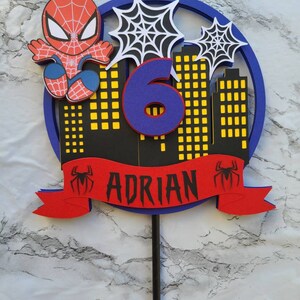 May include: A red and blue Spiderman cake topper with a blue number 6, a red banner with the name "ADRIAN" and a black spider web design.