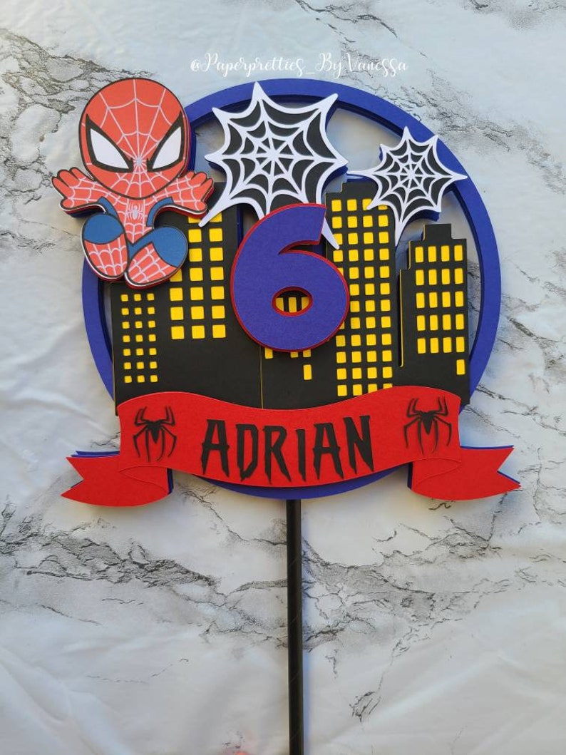 May include: A red and blue Spiderman cake topper with a blue number 6, a red banner with the name "ADRIAN" and a black spider web design.