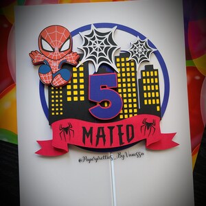 May include: A red and blue Spiderman cake topper featuring a black cityscape, a blue number 5, and a red banner with the name "MATED" in black letters. The topper is on a white background and includes the text "@Paperpretties_ByVanessa".