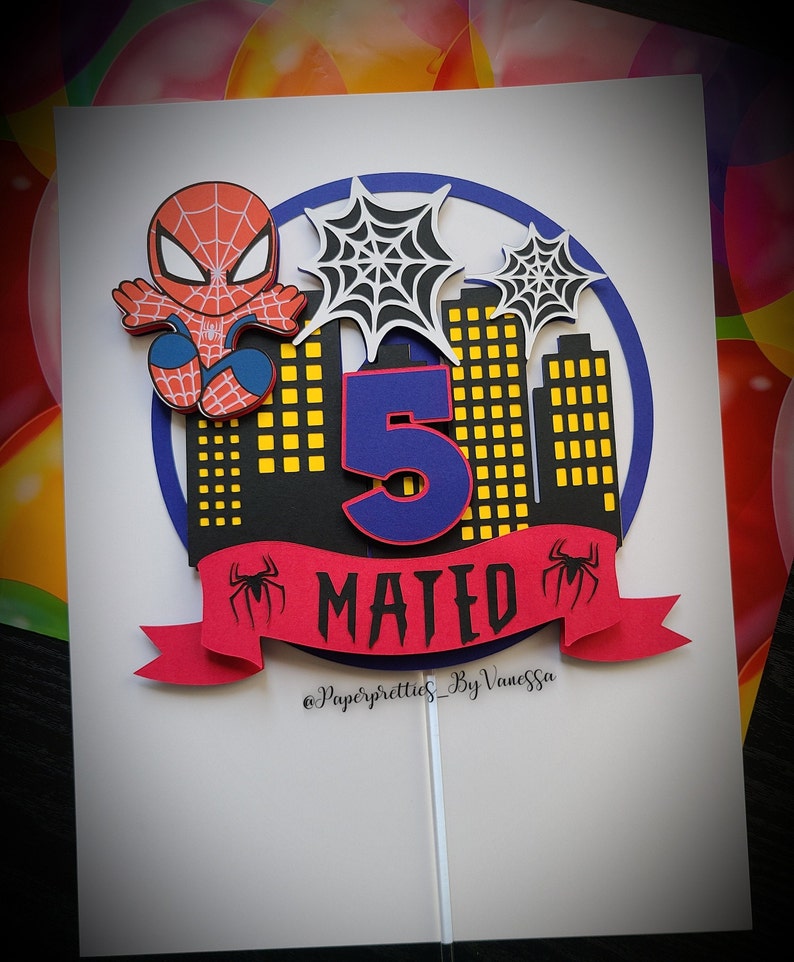 May include: A red and blue Spiderman cake topper featuring a black cityscape, a blue number 5, and a red banner with the name "MATED" in black letters. The topper is on a white background and includes the text "@Paperpretties_ByVanessa".