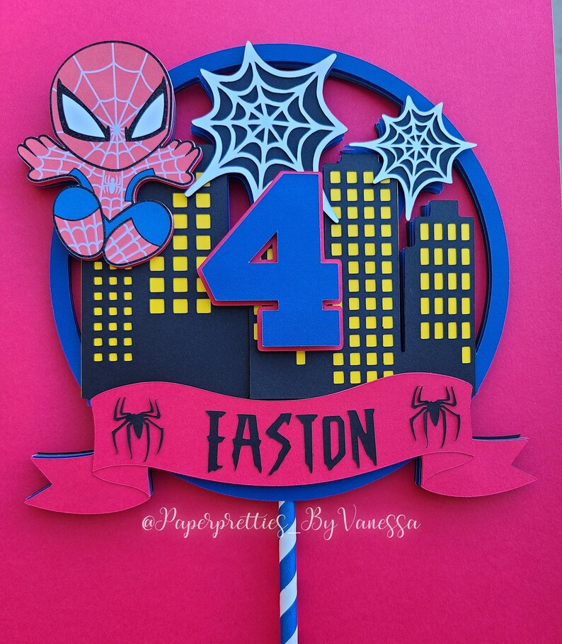 May include: A red and blue Spiderman cake topper with a number 4, a banner that says "Easton", and a cityscape in the background. The topper is on a stick with blue and white stripes.