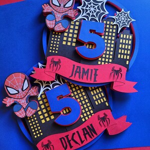 May include: Two Spiderman themed birthday cake toppers featuring red and blue Spiderman characters, a number 5, and a red banner with the names Jamie and Declan.