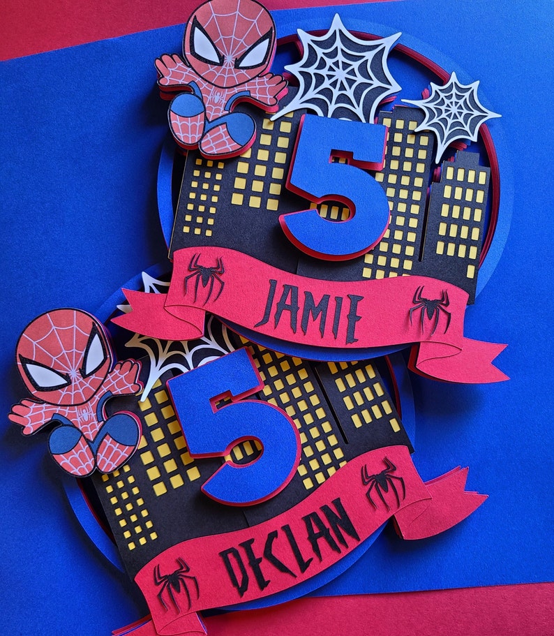 May include: Two Spiderman themed birthday cake toppers featuring red and blue Spiderman characters, a number 5, and a red banner with the names Jamie and Declan.
