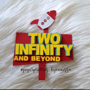 May include: Red and yellow paper sign with the text "TWO INFINITY AND BEYOND" and a white rocket ship with red accents on top. The sign has a white background with a red border. The text "@paperpretties_byanessa" is at the bottom of the sign.
