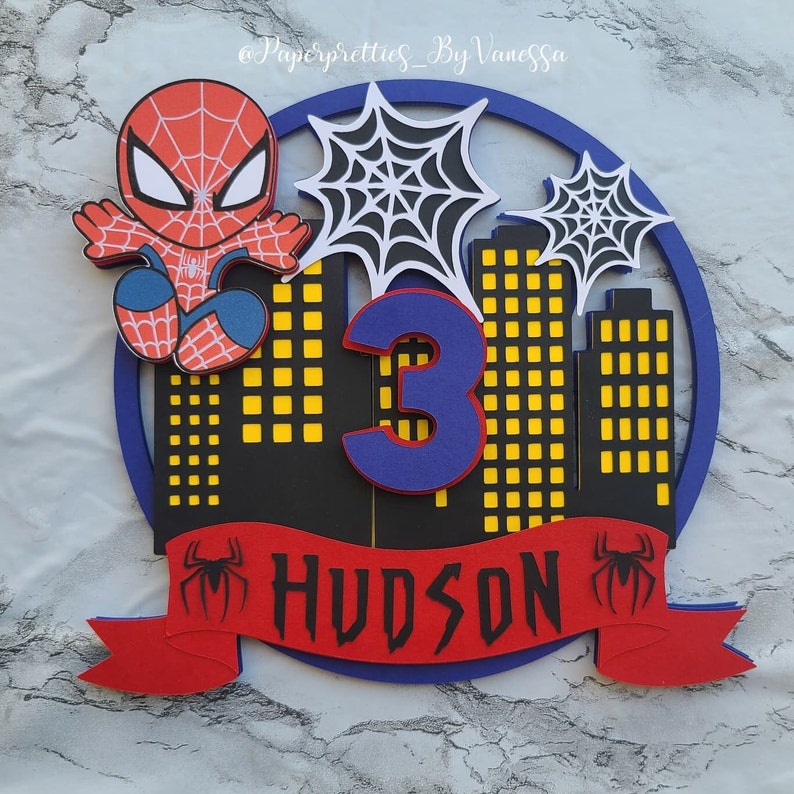 May include: A red and blue Spiderman cake topper with a red banner that says "HUDSON" and a number 3. The topper features a cityscape with yellow windows and two spiderwebs.