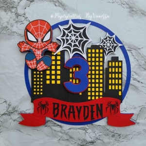 May include: A red and blue Spiderman cake topper with a black cityscape backdrop, a blue number 3, and a red banner with the name "Brayden" in black letters.