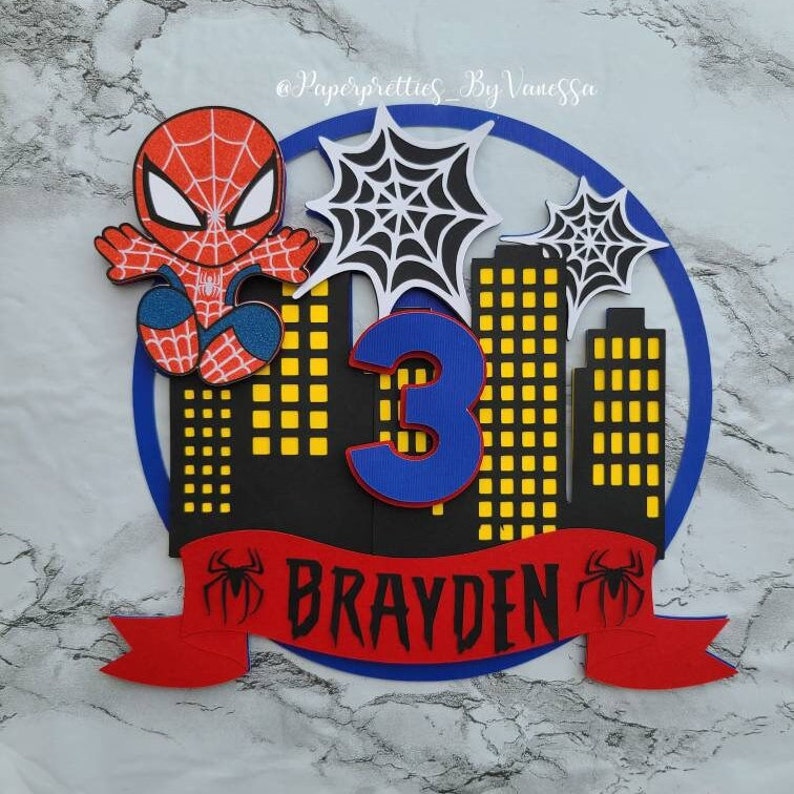 May include: A red and blue Spiderman cake topper with a black cityscape backdrop, a blue number 3, and a red banner with the name "Brayden" in black letters.