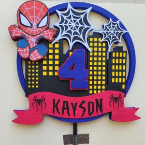 May include: A red and blue Spiderman cake topper with a red banner that says "KAYSON" and a blue number "4". The topper features a Spiderman character, a cityscape, and spiderwebs.