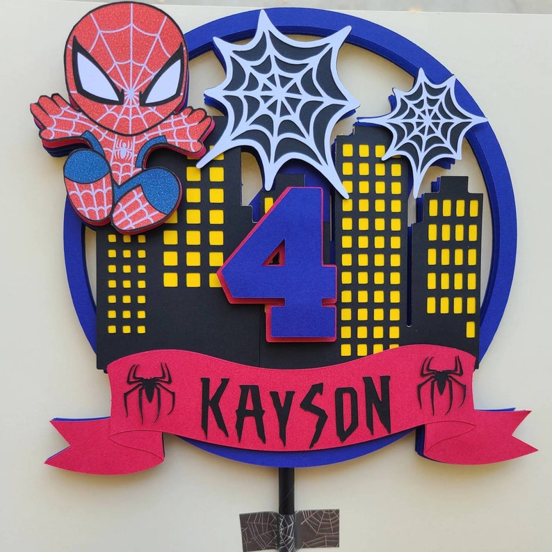 May include: A red and blue Spiderman cake topper with a red banner that says "KAYSON" and a blue number "4". The topper features a Spiderman character, a cityscape, and spiderwebs.