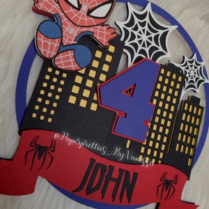 May include: A red, blue and black Spiderman themed cake topper featuring a Spiderman character, the number 4 and the name "JOHN" on a red banner. The background is a black cityscape with yellow windows.