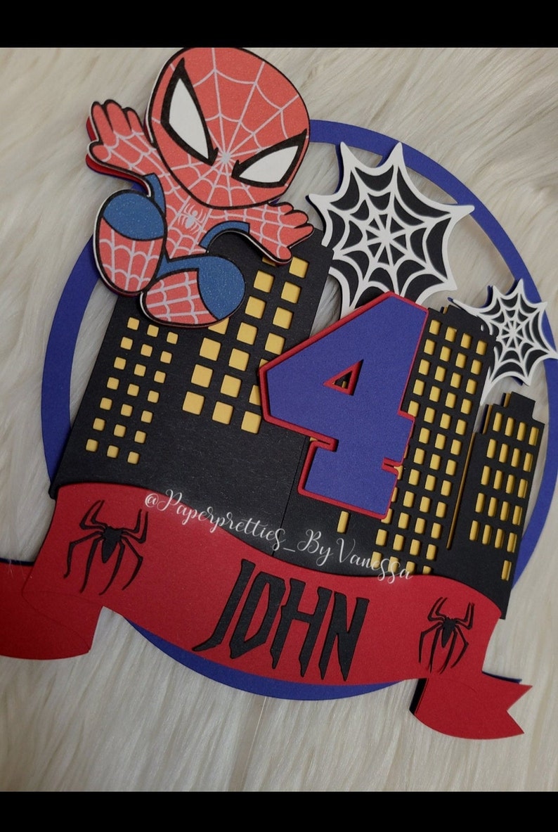 May include: A red, blue and black Spiderman themed cake topper featuring a Spiderman character, the number 4 and the name "JOHN" on a red banner. The background is a black cityscape with yellow windows.