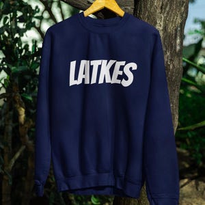 May include: A navy blue sweatshirt with the word "LATKES" in large white capital letters across the chest. The sweatshirt is hanging on a wooden hanger against a natural background of trees and foliage.