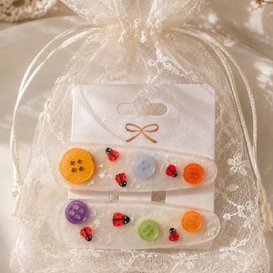 May include: Two clear resin hair clips featuring embedded colourful plastic buttons and small red ladybug charms. The clips are displayed on a white cardboard backing card inside a sheer white lace drawstring pouch with a delicate floral pattern.