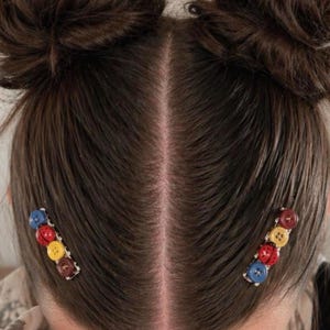 May include: Two hair clips featuring a row of four colorful buttons each, including red, yellow, blue, and dark brown circular buttons. The clips are attached to dark brown hair styled in two buns on either side of a center part, securing the hair in place.